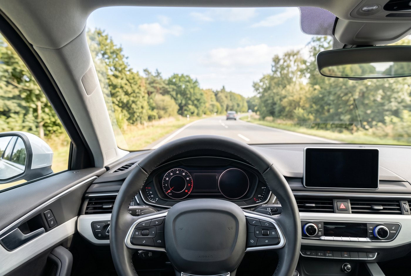 Bright view over a clean steering wheel toward a sunlit tree-lined road